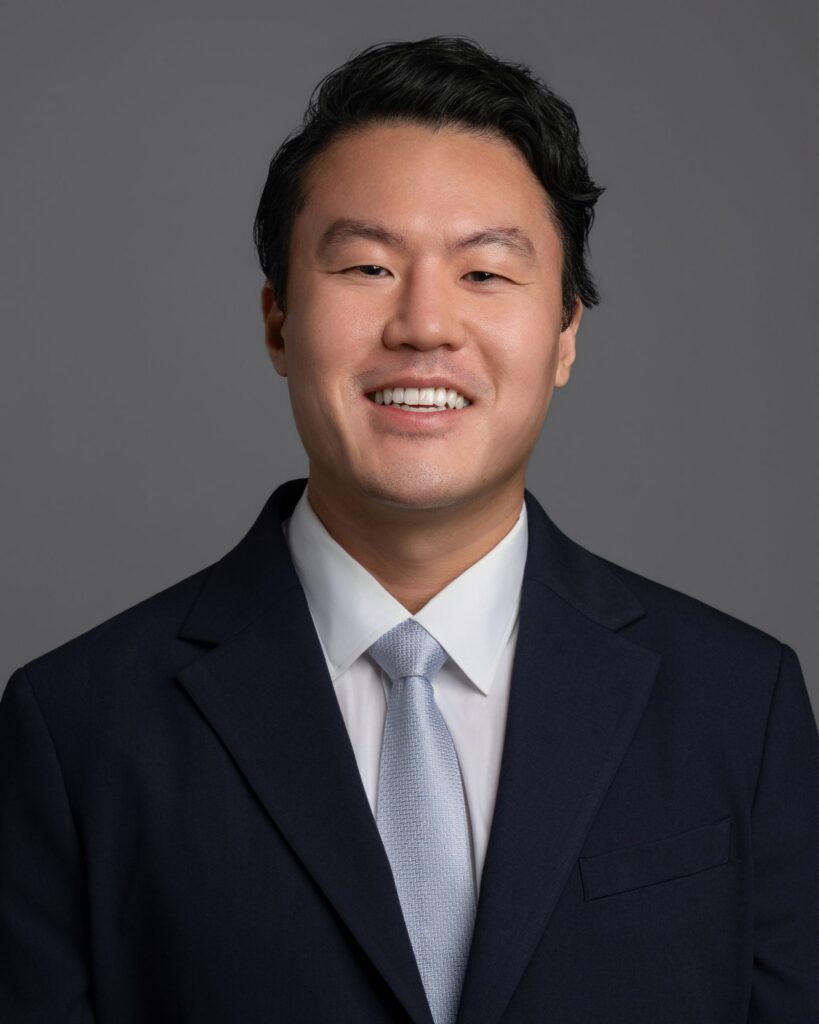 Smiling man in a dark suit and light blue tie, portrait against a gray background, cropped at shoulders.